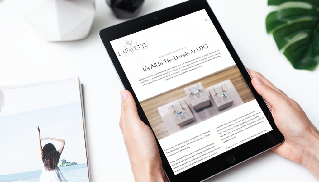 A person holds a tablet displaying a custom website design for a jewelry store featuring three rings on stands. A magazine with a woman raising her arms and a green plant are partially visible on the white table in New York.