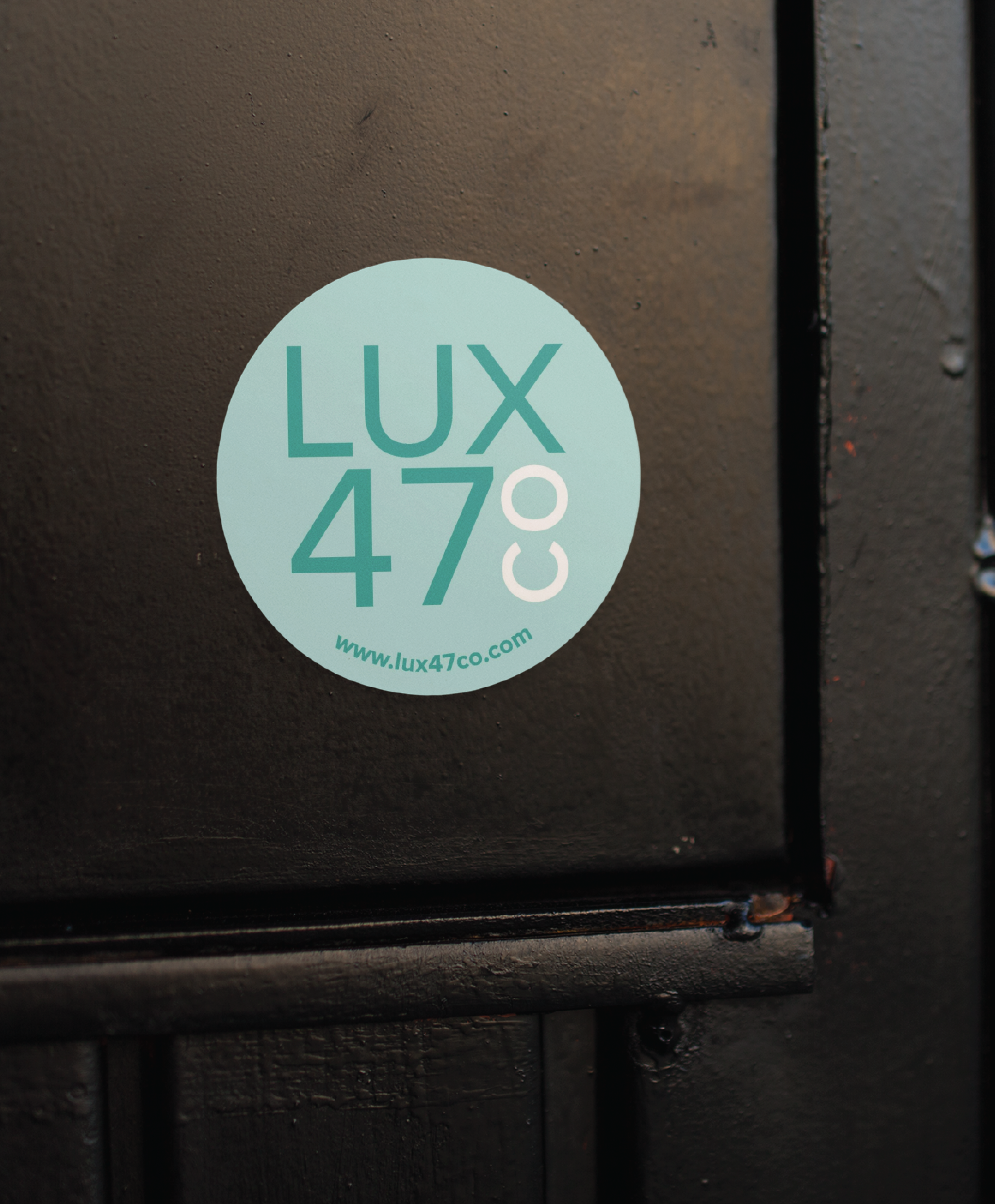 A teal circular sticker featuring LUX 47 CO, www.lux47co.com, and a stylish print design is displayed on a dark-colored, textured surface, possibly a door or wall.