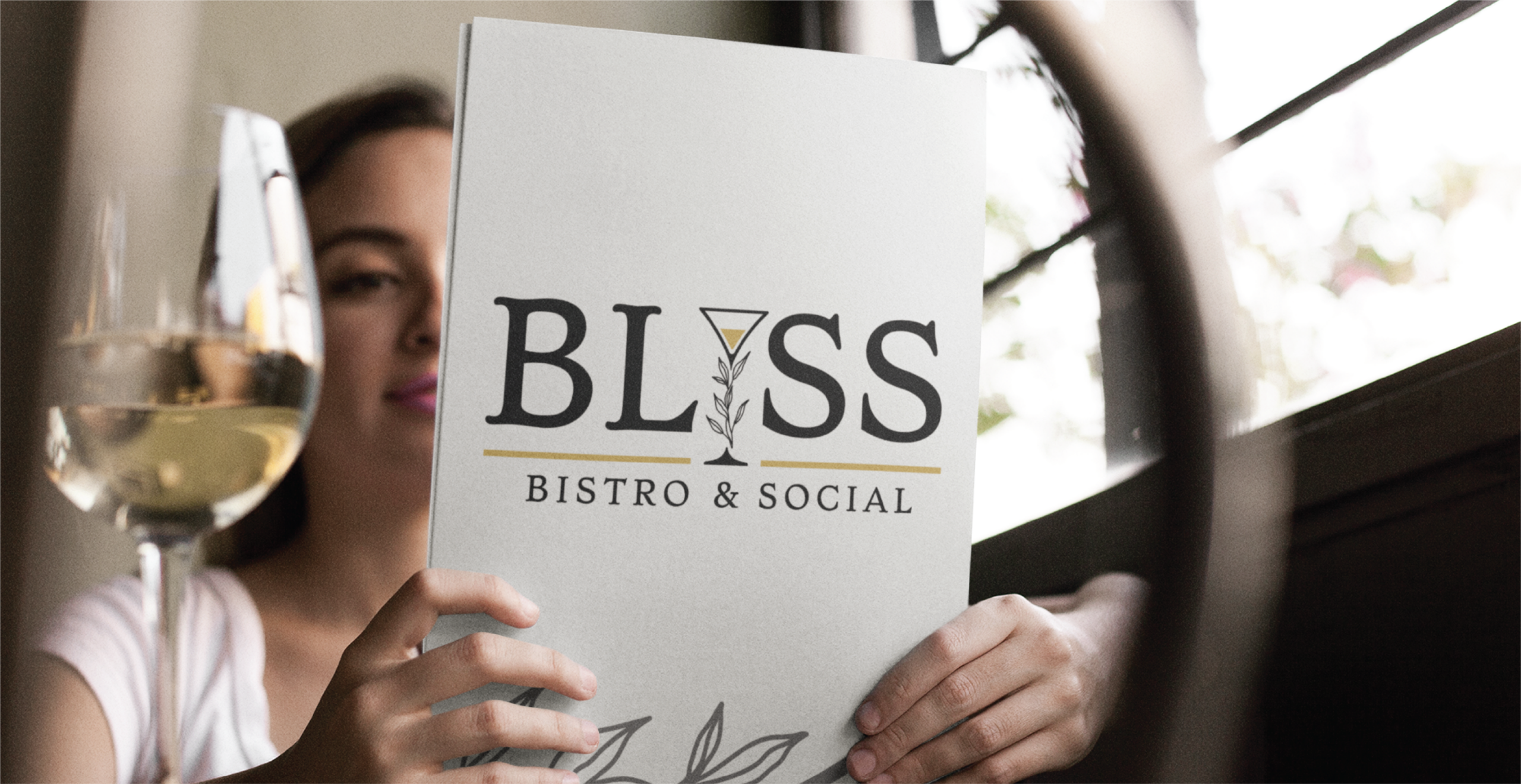 A person holds a menu that reads “BLISS BISTRO & SOCIAL” with a wine glass in the foreground, sitting by a window with natural light at this Florida restaurant known for its custom logo design.
