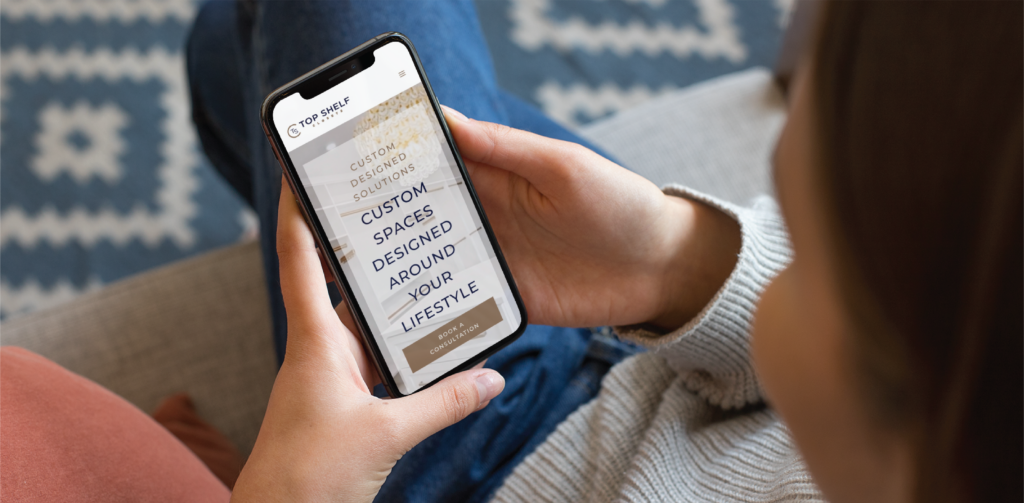 A person holds a smartphone displaying a website with the text Custom spaces designed around your lifestyle, while sitting on a couch in Pennsylvania, with part of a blue-patterned rug visible in the background.