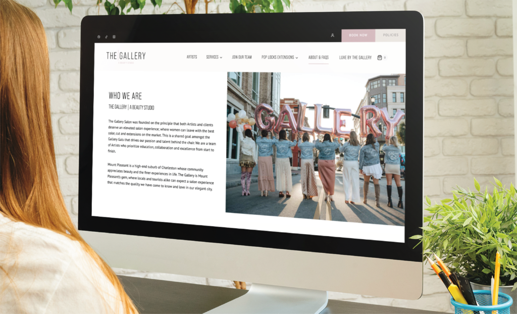 A person views a beautifully crafted website design for an art gallery on a desktop computer. The screen displays a section titled “Who We Are” with text on the left and an image of people holding balloon letters spelling GALLERY on the right.