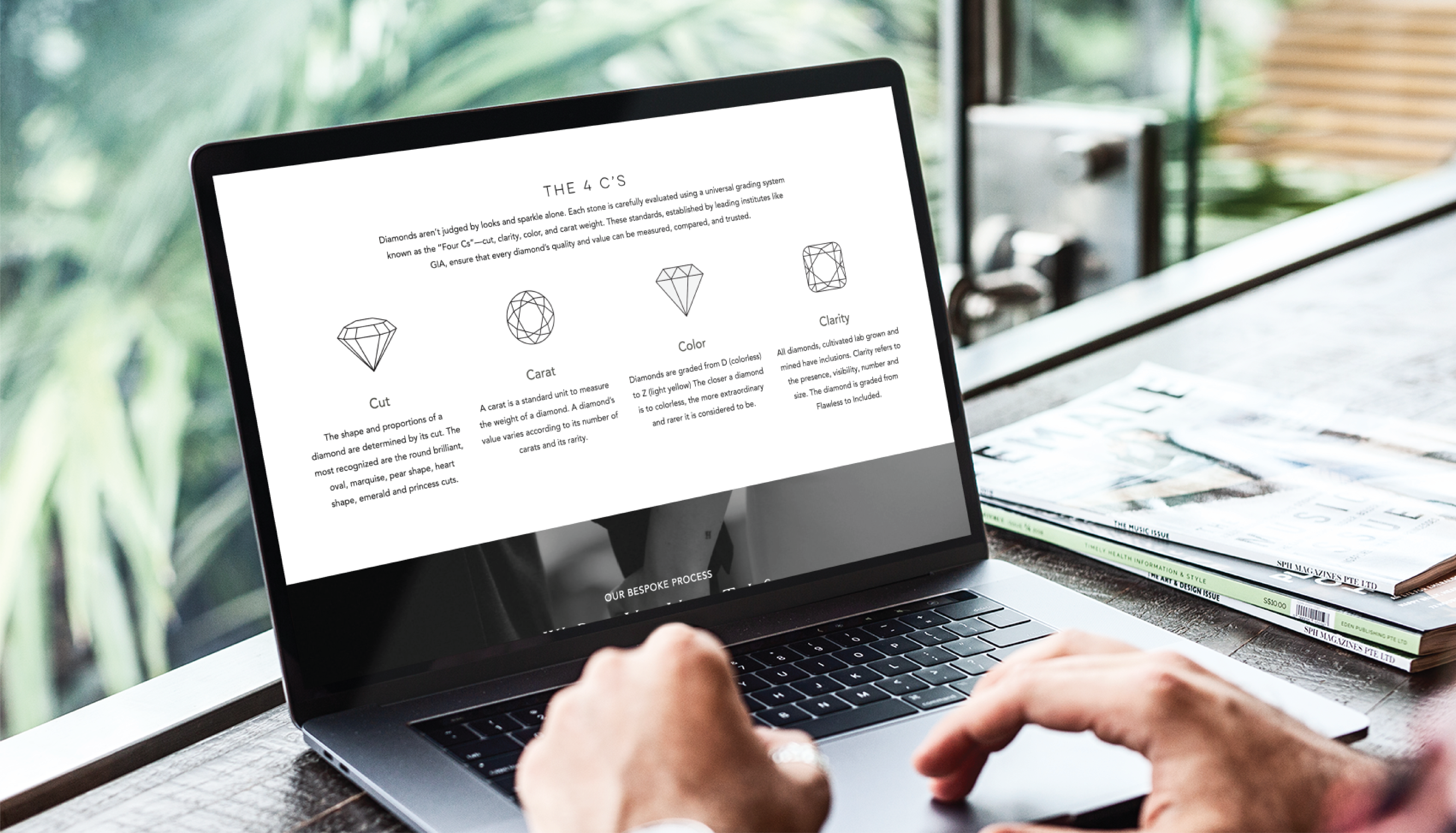 A person uses a laptop displaying an educational page about the 4 C’s of diamonds—Cut, Carat, Color, and Clarity—crafted through custom website design, alongside stacked magazines on a desk by a window.