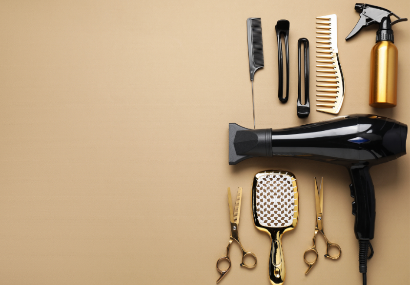 A flat lay of hairdressing tools on a beige background, perfect for a salon website, featuring a black hairdryer, golden spray bottle, combs, brushes, hair clips, and scissors neatly arranged on the right side.