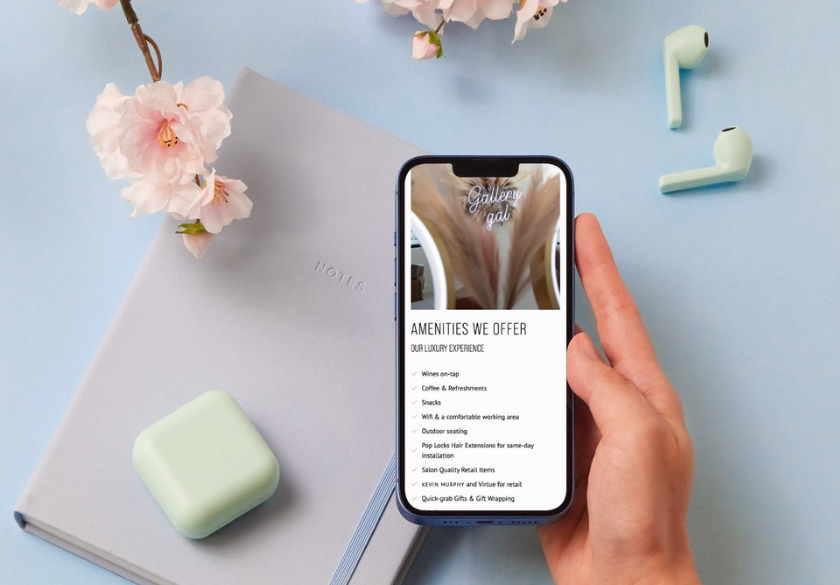 Hand holding a smartphone showing a flyer titled Amenities We Offer, on a blue desk with a notebook and pastel mint earbuds case.