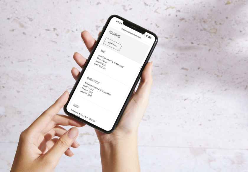 Hands holding a smartphone showing a web page with a 'COLORING' section and a 'BOOK NOW' button.