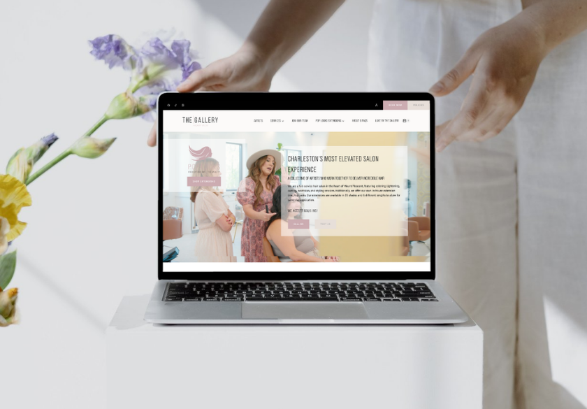 A person holds a laptop displaying a Hair Salon Website called The Gallery, featuring three women inside a salon. A hand holds the laptop, with pastel flowers in the background, highlighting modern Hair Salon Website Design.