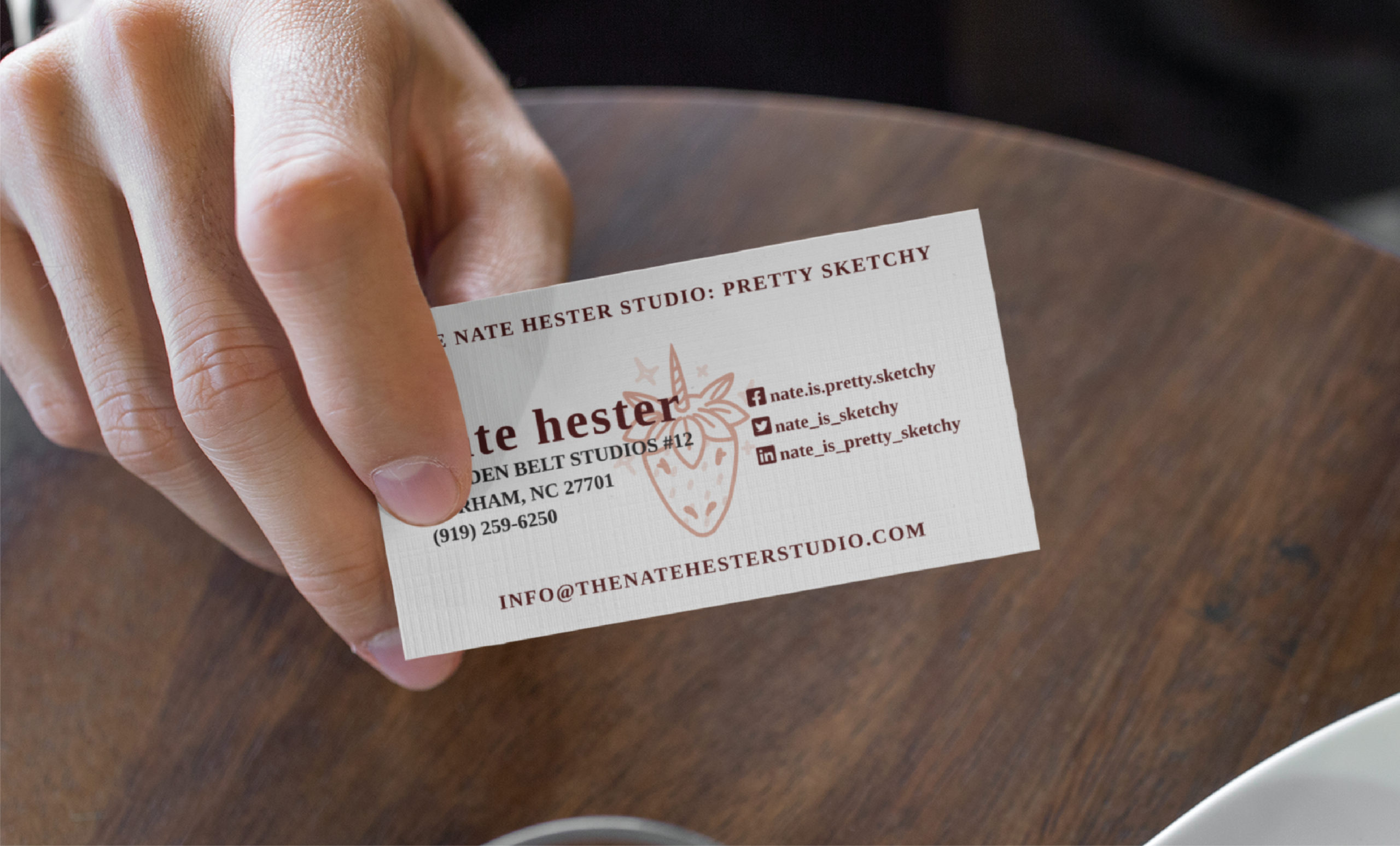 A person holds a business card for Nate Hester Studio: Pretty Sketchy, a Tennessee-based business card creation service featuring contact details, a fox logo, and social media handles, over a wooden table.
