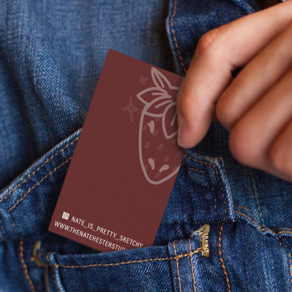 A hand tucks a maroon business card featuring an artist's strawberry illustration, Instagram handle, and website into the pocket of blue denim jeans—showcasing creative print design from North Carolina.