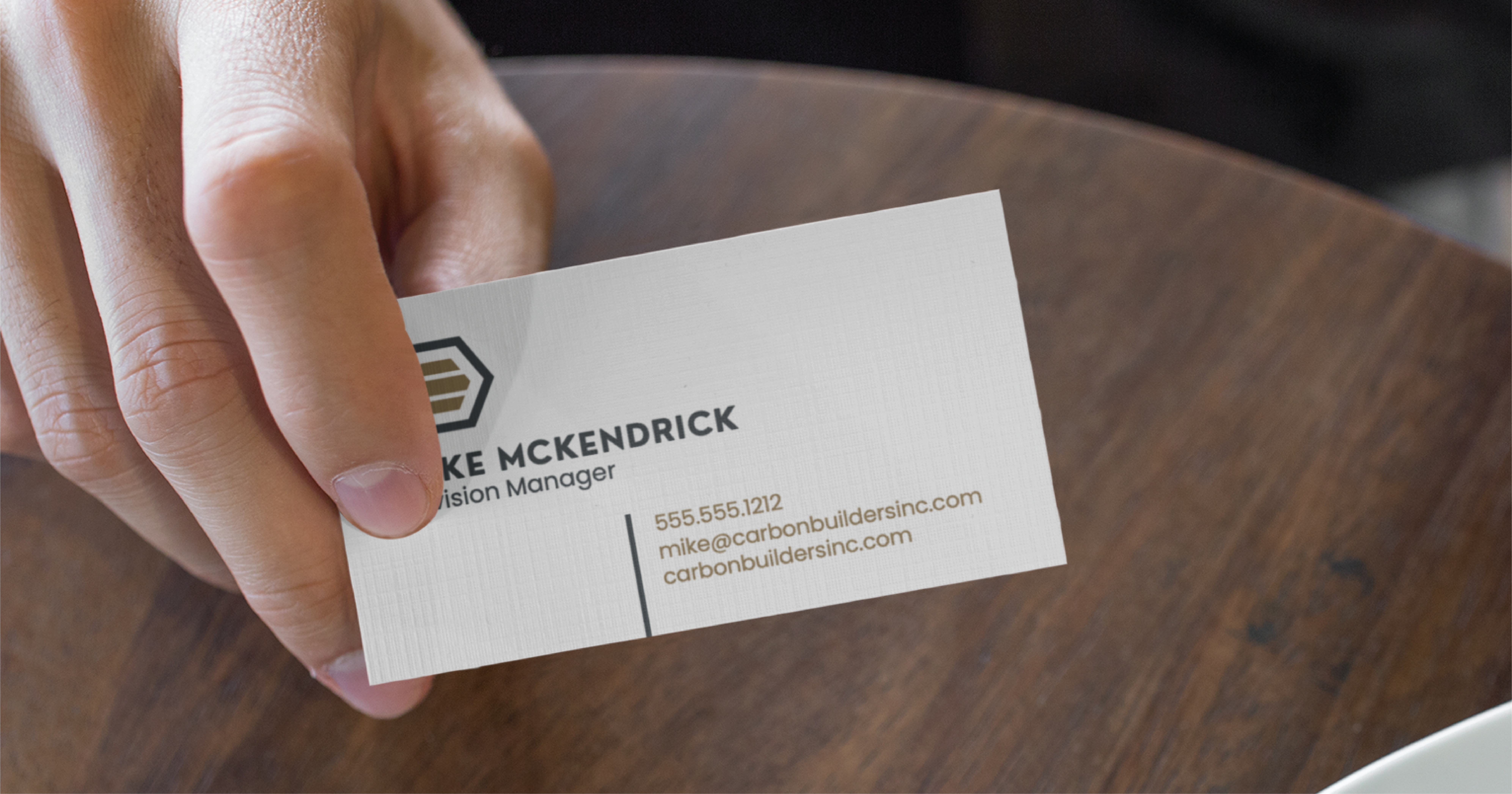 A hand holding a business card for Mike McKendrick, Division Manager at Carbon Builders Inc., showcasing expert print design, his email, phone number, and the company’s website on a wooden table background in North Carolina.