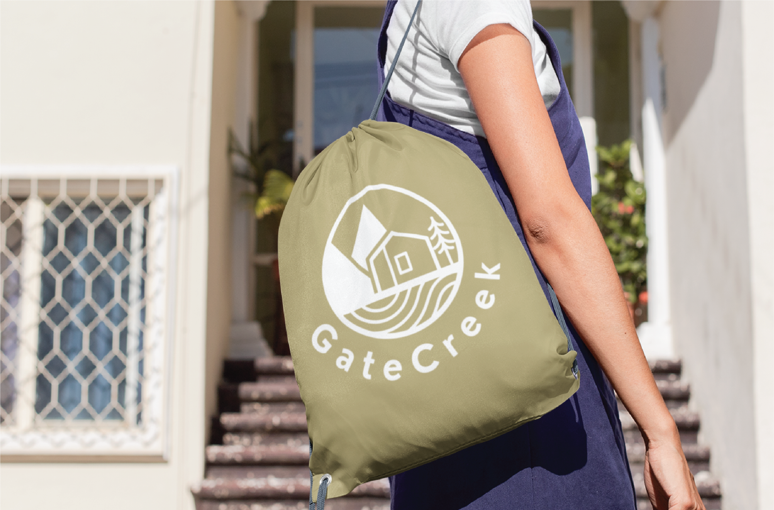 A person stands outside near steps, carrying an olive green drawstring bag with a white logo design featuring a cabin, trees, and the words GateCreek—perfect for a women-focused business in the US.