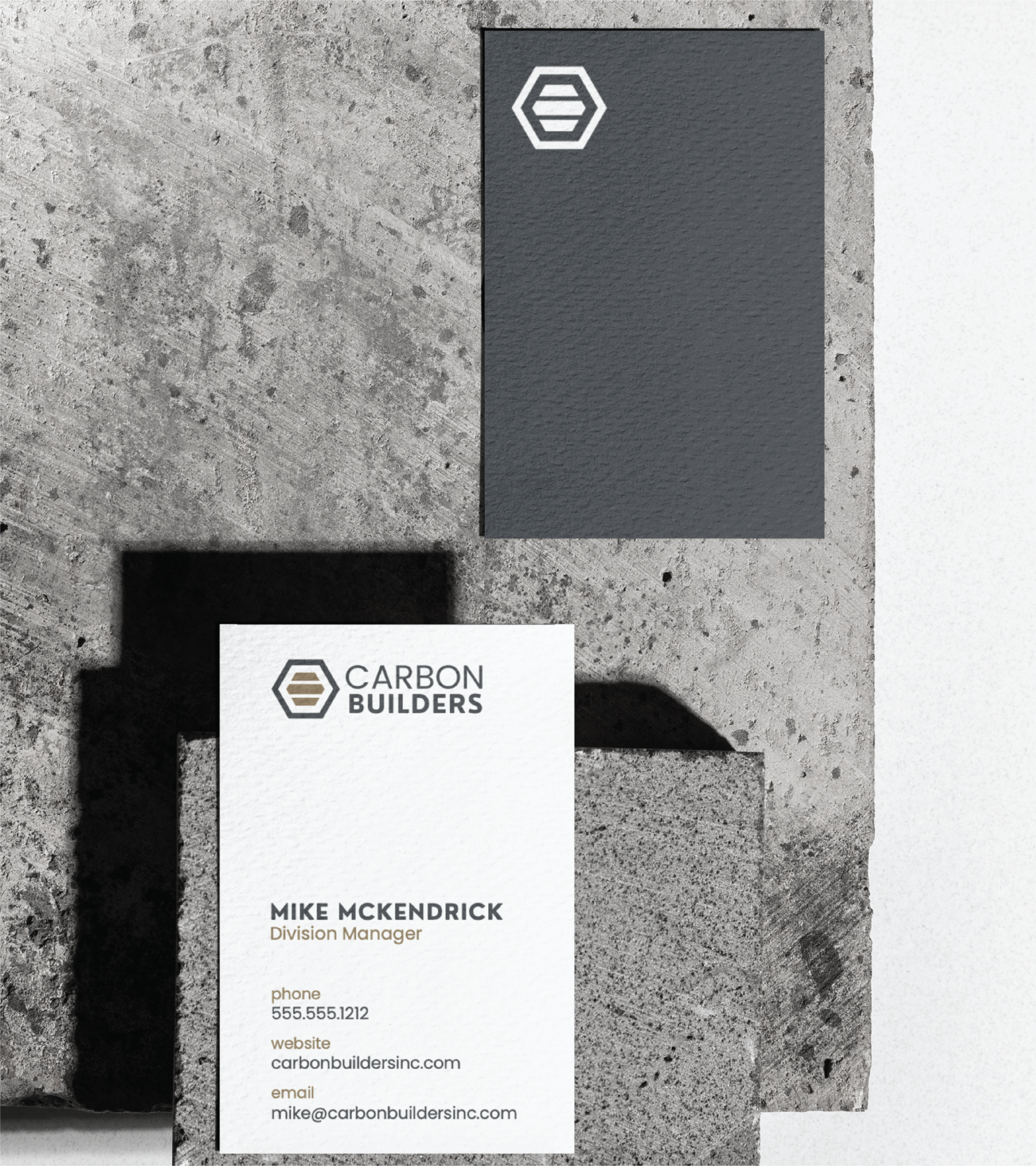 A business card for Carbon Builders, a Michigan General Contractor, with a hexagon logo, rests on a textured concrete surface. The custom stationery lists contact details for Mike McKendrick, Division Manager, plus the company's website and email.