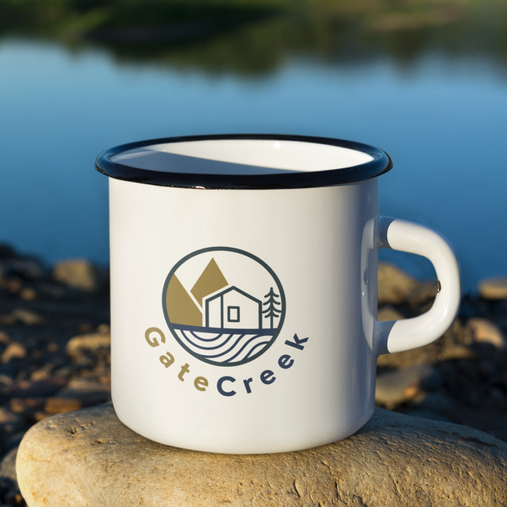 A white enamel mug with a black rim sits on a rock by the water. The mug features a Property Group logo design with a cabin, trees, mountains, and the text “GateCreek” beneath the design.