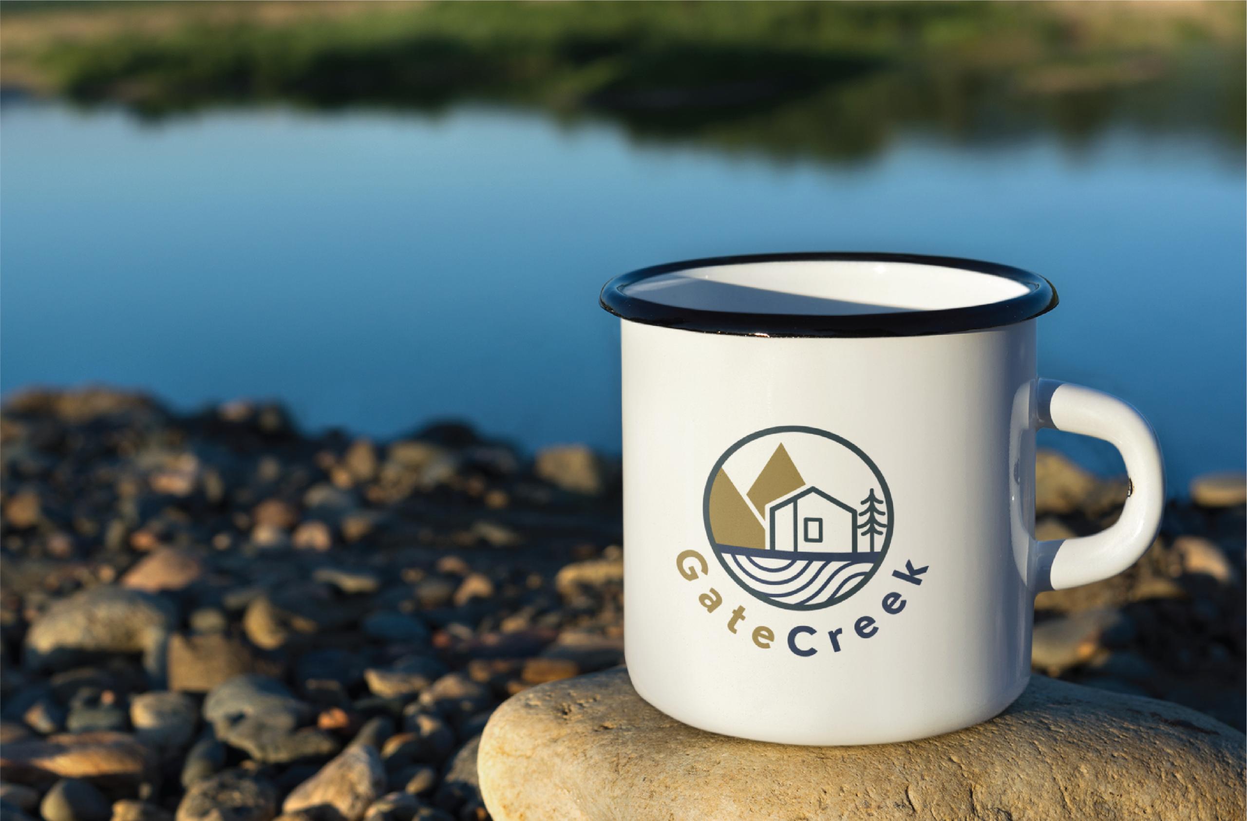 A white enamel mug with a women-focused business logo design, featuring GateCreek and a cabin, sits on a rock by a US riverbank, with smooth stones and calm blue water in the background.