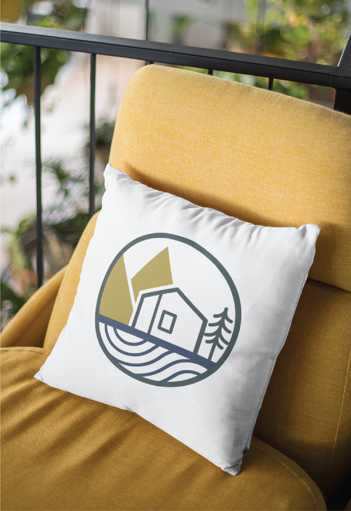 A white throw pillow featuring a minimalist logo design of a house, tree, and waves in a circular frame rests on a mustard yellow chair, with a blurred background of green plants and railing—ideal for any US women-focused business space.