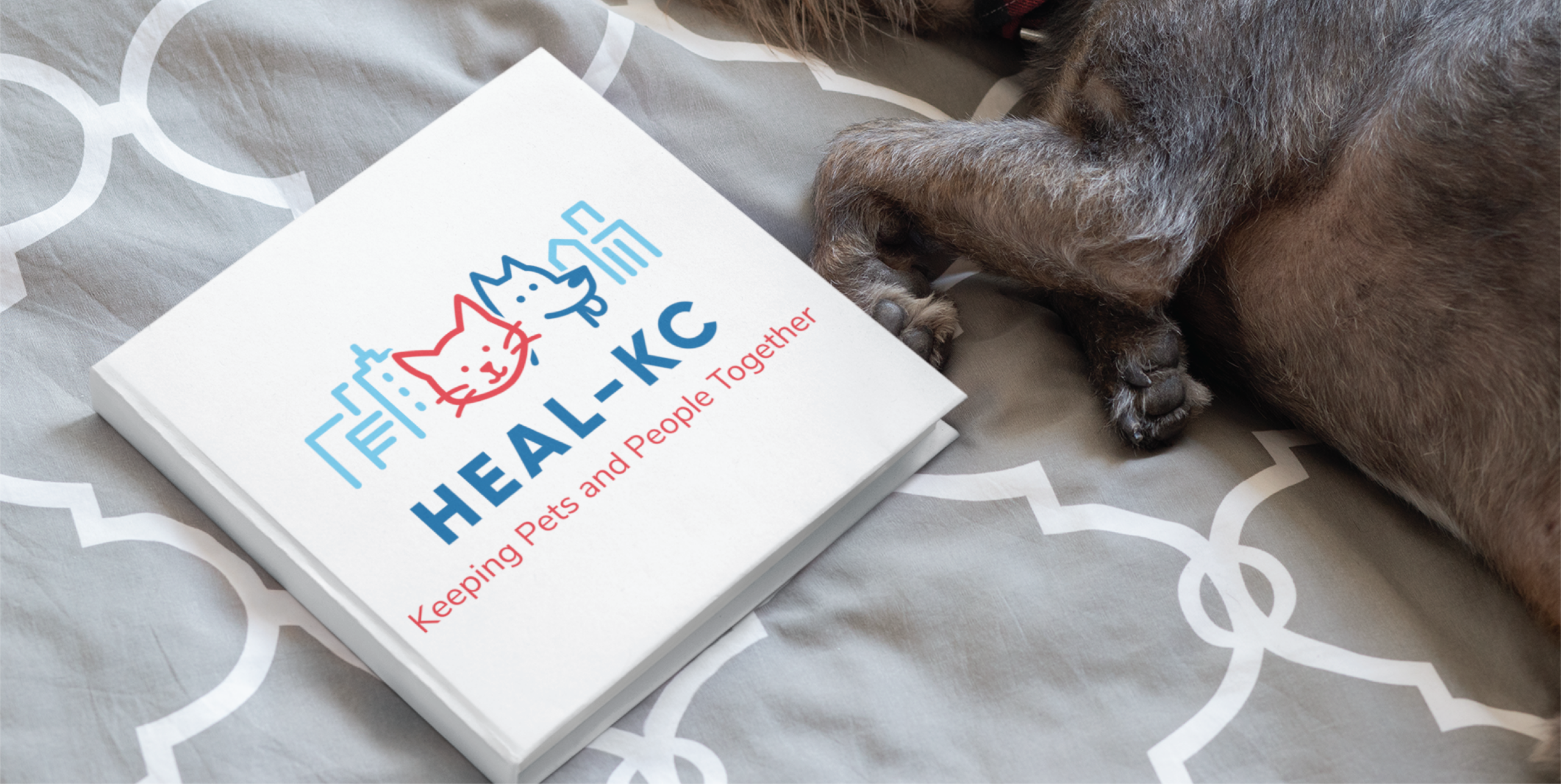 White booklet with a blue/red logo (dog and cat) and the text 'Keeping Pets and People Together' on a light bedspread; a dog's paw rests nearby on the right side.