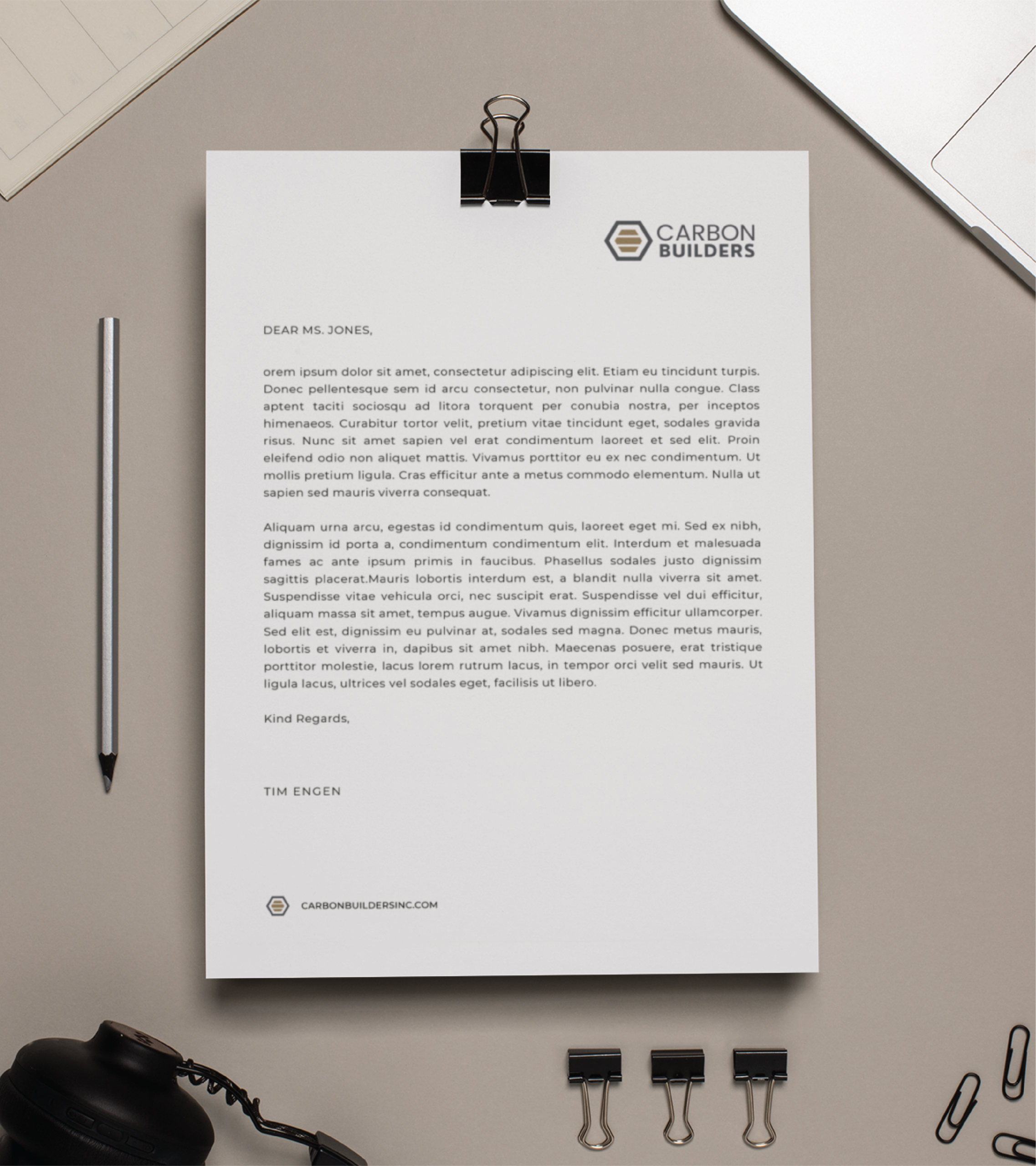 A letter on CARBON BUILDERS stationery—highlighting their role as a Michigan general contractor—rests on a desk with a pen, headphones, binder clips, and laptop keyboard. Addressed to Ms. Jones and signed by Tim Engen.