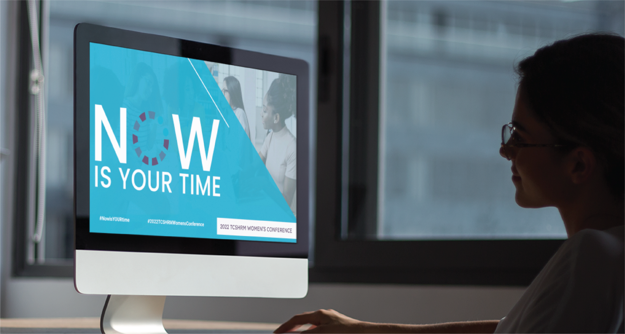 A person sits at a desk in Michigan, looking at a computer screen displaying “NOW IS YOUR TIME” alongside conference-related hashtags and social media graphics, with a faint image of people in discussion nearby.