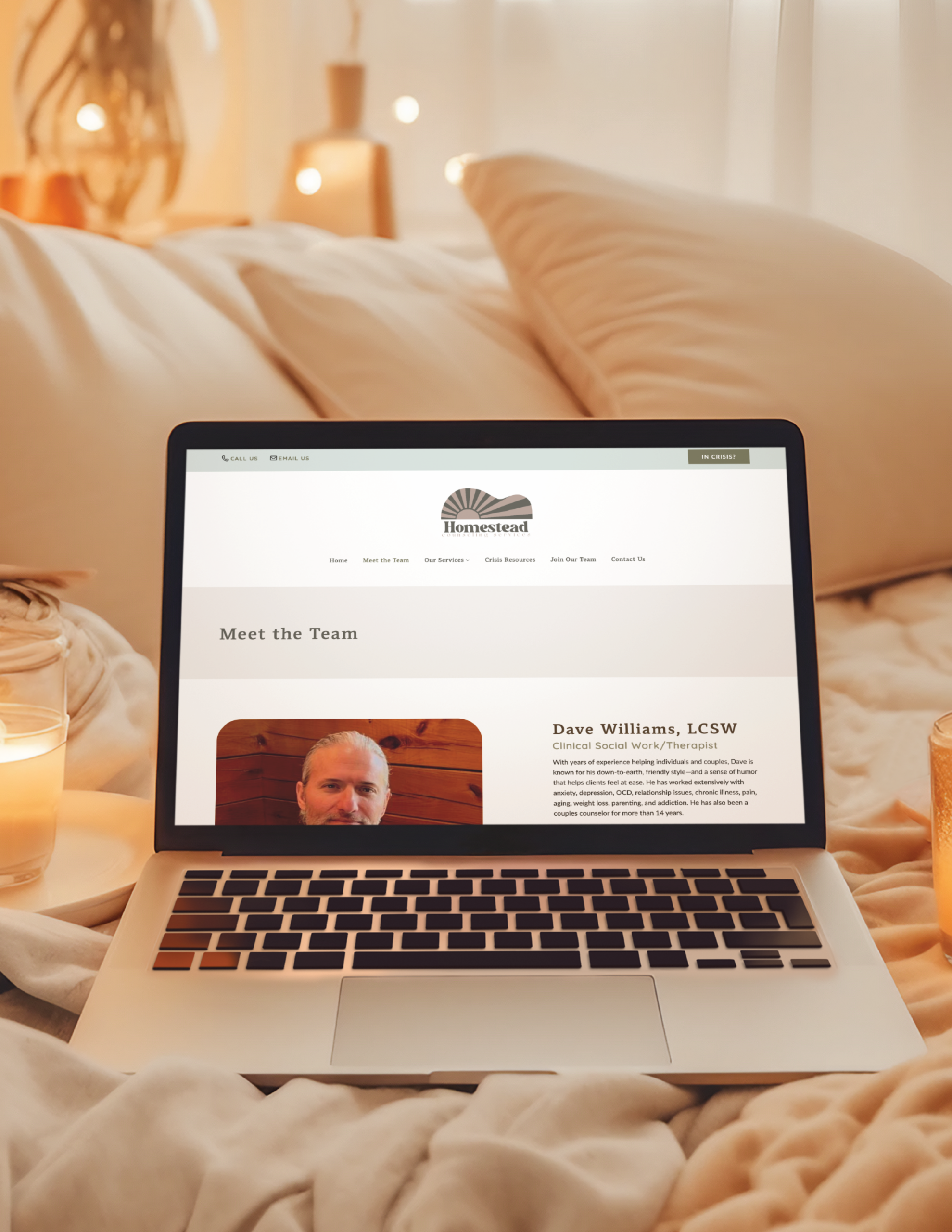 A laptop rests on a cozy bed near lit candles, displaying Homestead Counseling’s Meet the Team webpage. Showcasing thoughtful website design, it features a photo and bio of bearded Dave Williams, LCSW. Warm lighting enhances the inviting Pennsylvania setting.