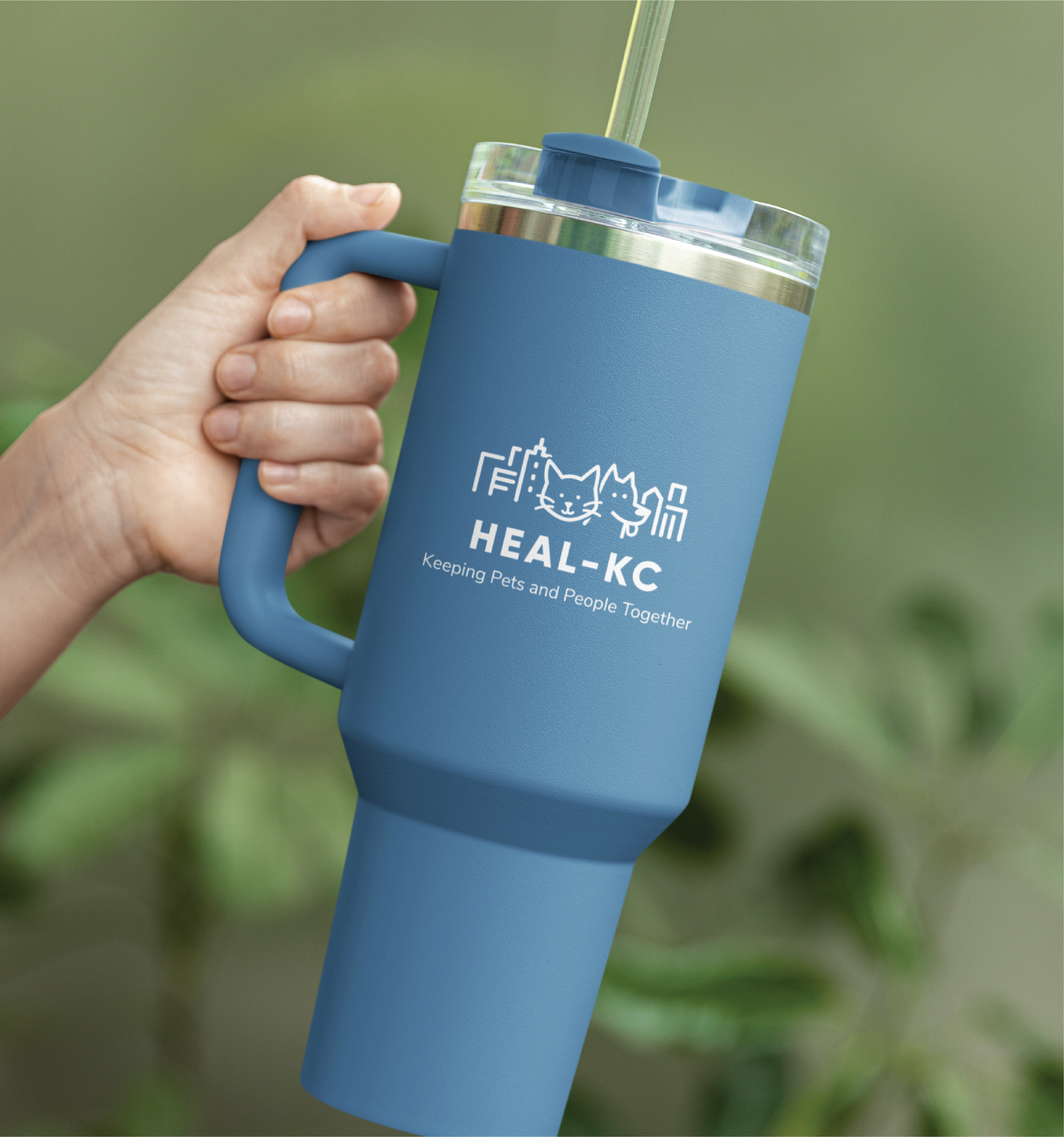 A hand holds a large blue tumbler with a clear lid and straw, featuring a city skyline, dog, cat, and the “HEAL•KC Keeping Pets and People Together” logo design. Greenery is blurred in the background, giving it a fresh US vibe.