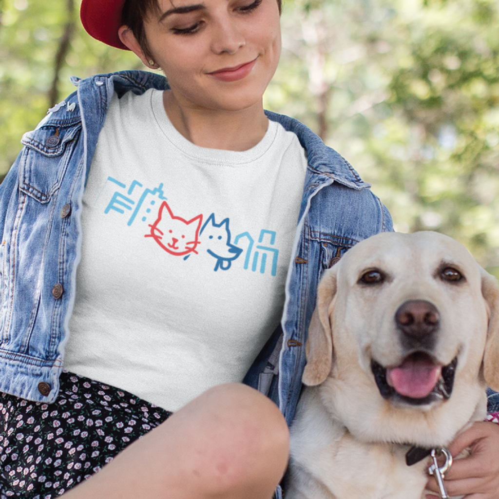 A woman wearing a red hat and denim jacket smiles while sitting outdoors with her happy yellow Labrador. Her t-shirt features cartoon cat and dog faces with a Kansas City cityscape design in blue and red. Trees are in the background.