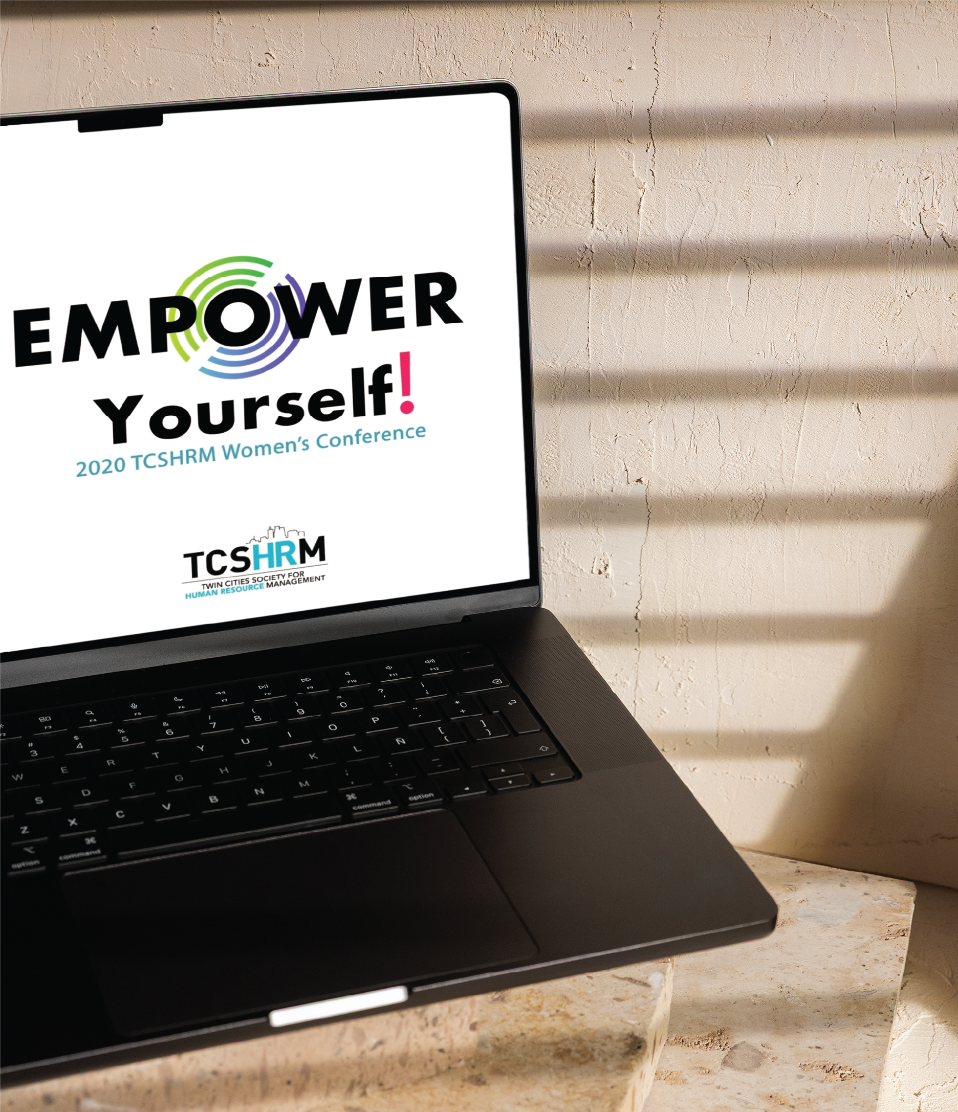 A laptop displays a screen with the text EMPOWER Yourself! 2020 TCSHRM Womens Conference and the TCSHRM logo. Sunlight casts shadows on the beige wall and table, while social media graphics from Michigan are highlighted on screen.