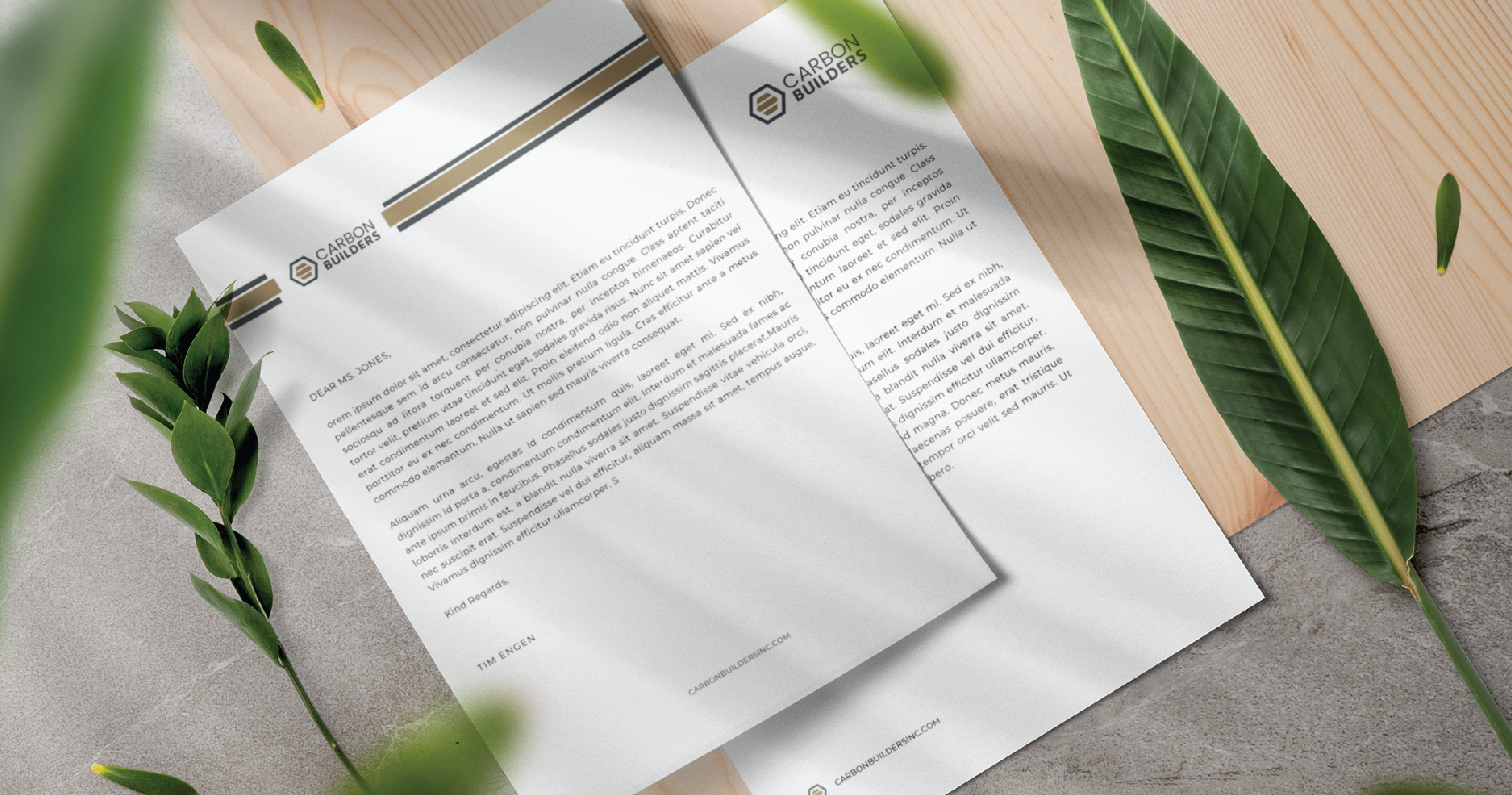 Two sheets of letterhead paper featuring Carbon Builders’ logo and text showcase a refined stationery design, lying on a gray surface beside green tropical leaves and a wooden board—perfect for a Michigan general contractor’s professional touch.