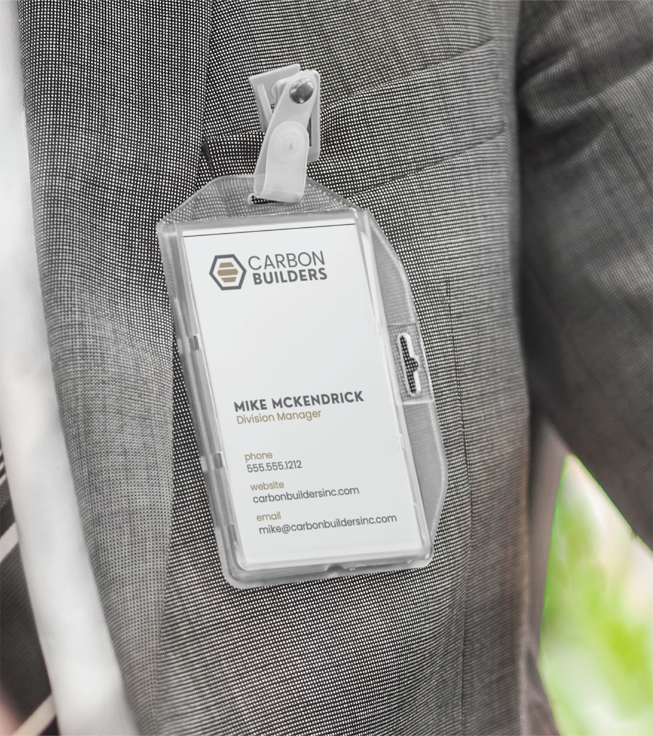 A close-up of a person wearing a gray suit with a name badge clipped to their jacket. The badge displays Carbon Builders, a Michigan General Contractor, and contact info for Mike McKendrick, Division Manager.