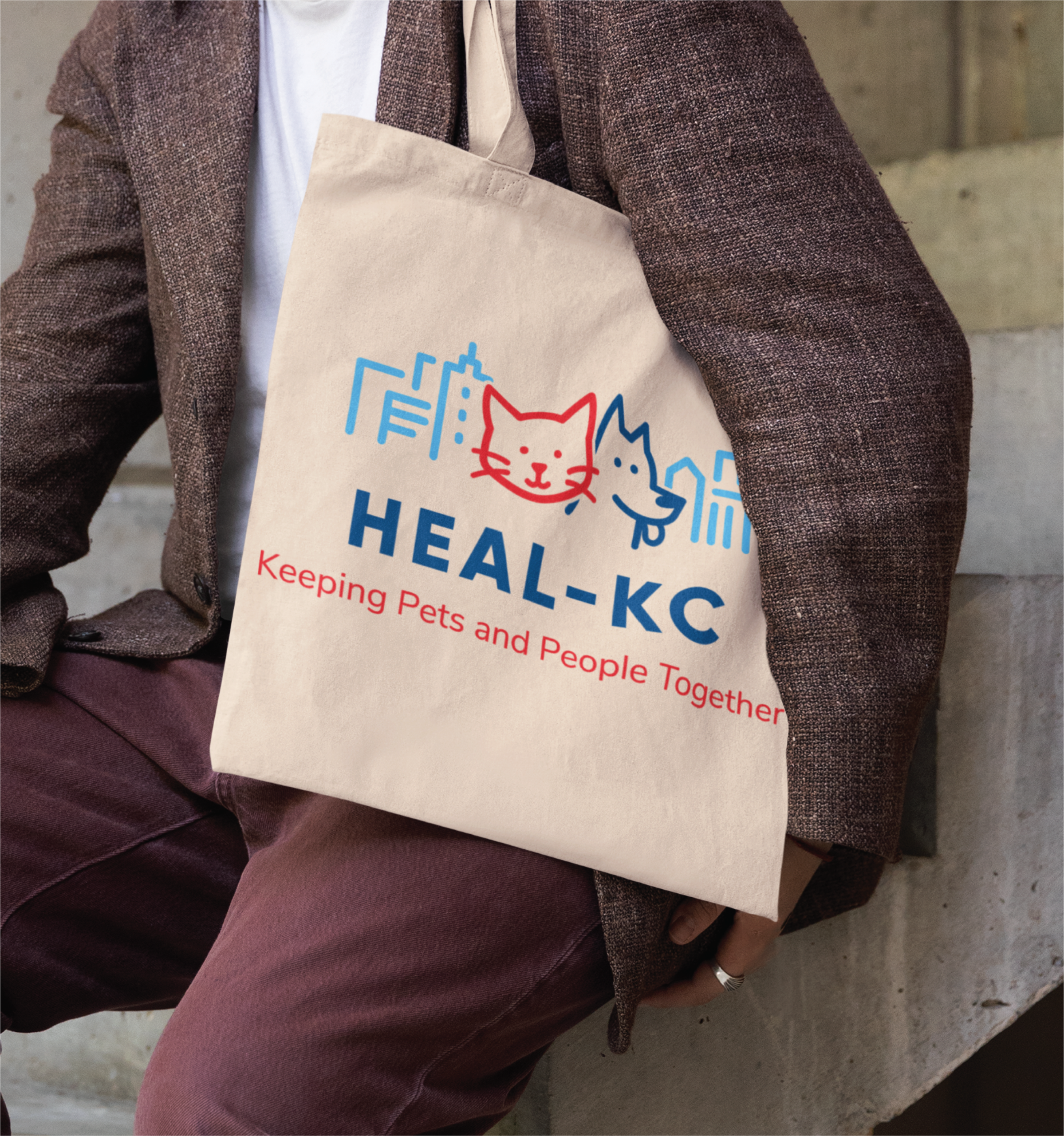 A person holds a beige tote bag with colorful illustrations of a cat and dog, city skyline, and the text “HEAL-KC Keeping Pets and People Together” printed on the front, showcasing creative US logo design.