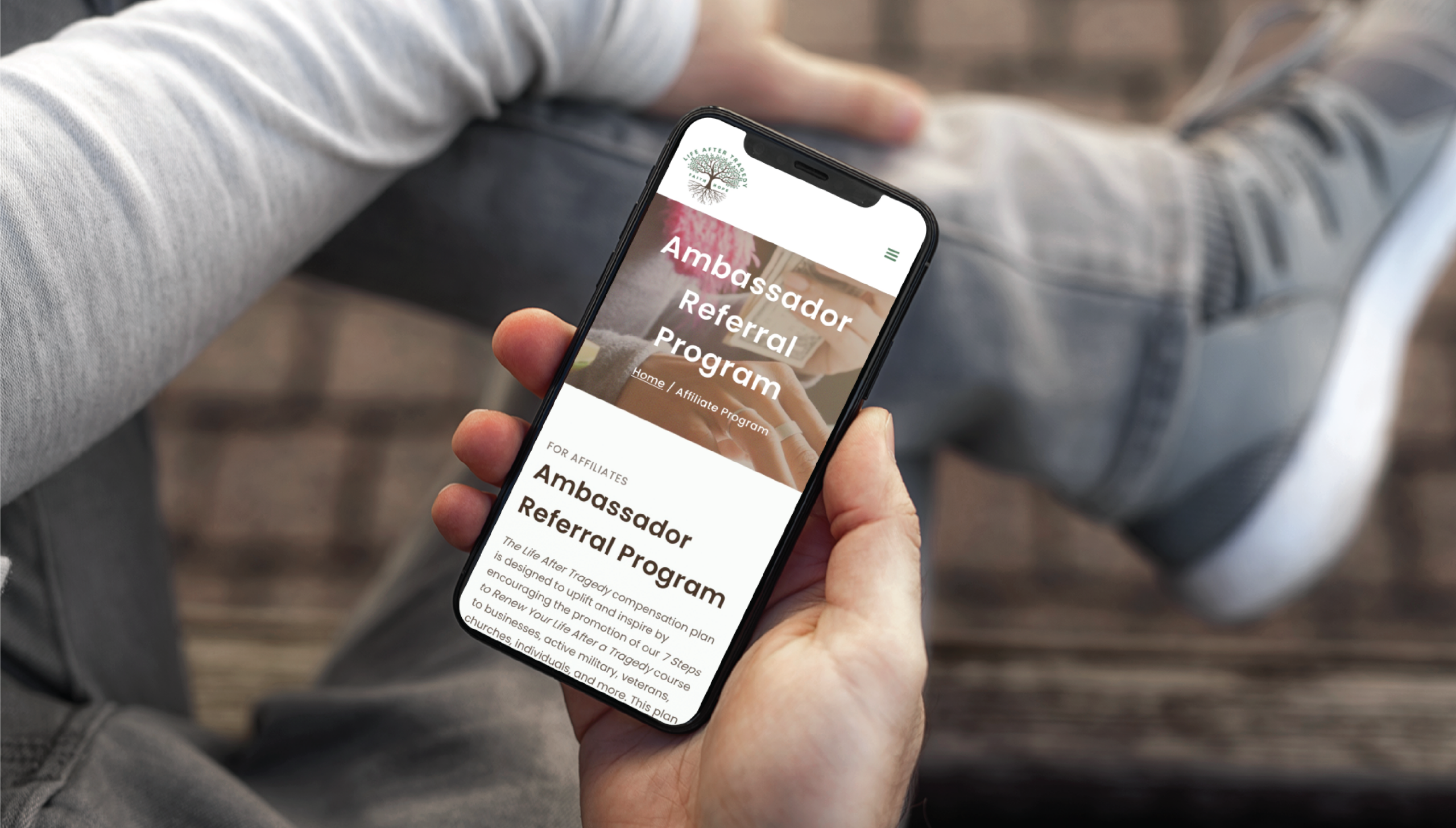 A person holds a smartphone displaying a website titled Ambassador Referral Program, featuring custom website design. The screen shows text and a blurred background while the person, wearing jeans and sneakers, sits cross-legged—perhaps in sunny Florida.