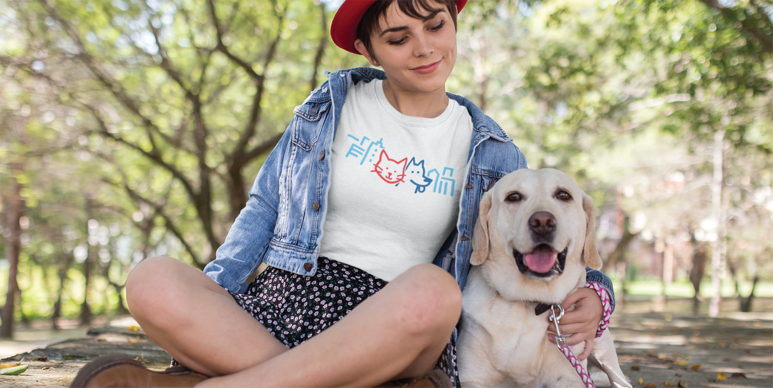A woman in a red hat and denim jacket sits on grass beside a happy yellow Labrador. She smiles, wearing a white t-shirt featuring a playful cat and dog logo design in blue and orange. Trees and sunlight fill the background.