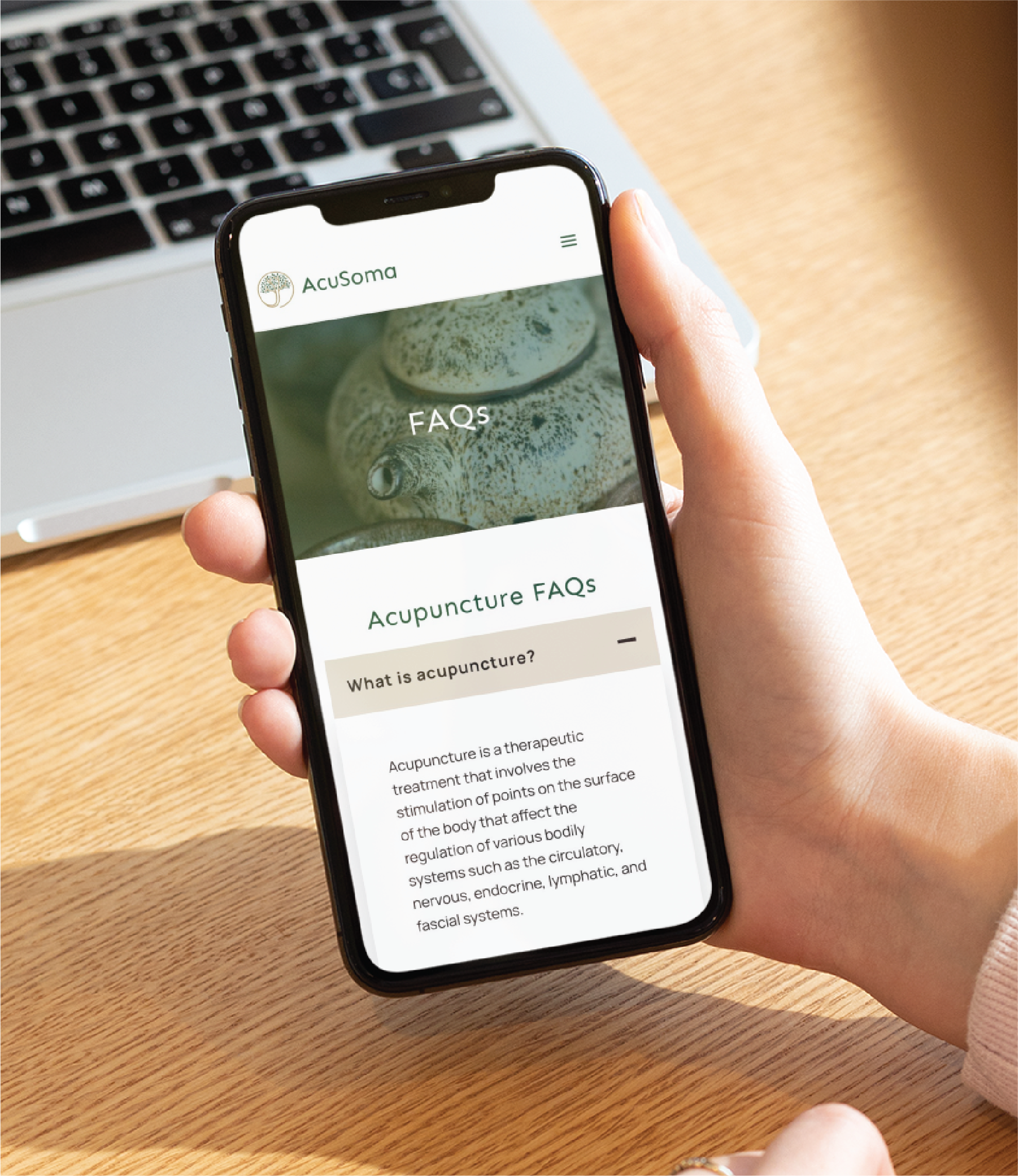 Hand holding a smartphone showing the AcuSoma website with acupuncture FAQs over a wooden desk and laptop keyboard in the background.