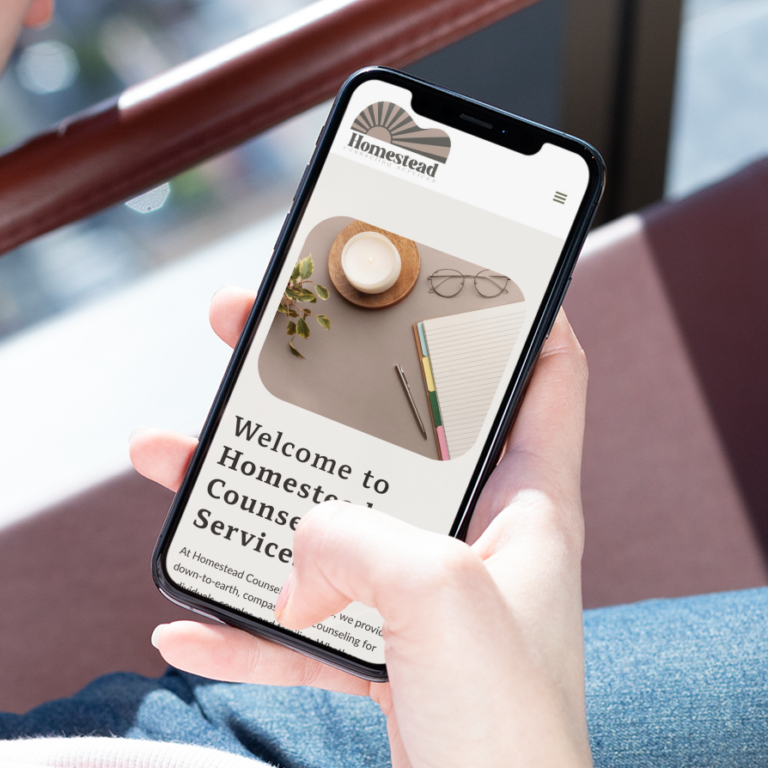 A person holds a smartphone displaying the Homestead Counseling Services website, highlighting its calming website design with a candle, plant, and stationery on a desk—perfect for anyone seeking counseling service in Pennsylvania.