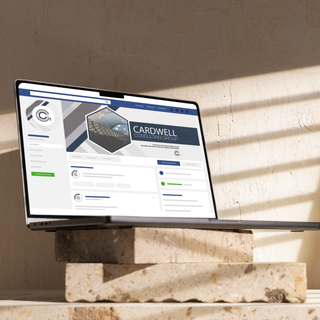 A laptop displaying a business Facebook page for Cardwell Consulting Group sits on stacked stone tiles in sunlight, with shadows from window blinds cast on the wall behind—highlighting their expertise in Social Graphics for Michigan businesses.