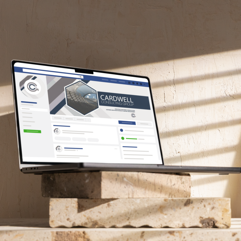 A laptop displaying a business Facebook page for Cardwell Consulting Group sits on stacked stone tiles in sunlight, with shadows from window blinds cast on the wall behind—highlighting their expertise in Social Graphics for Michigan businesses.
