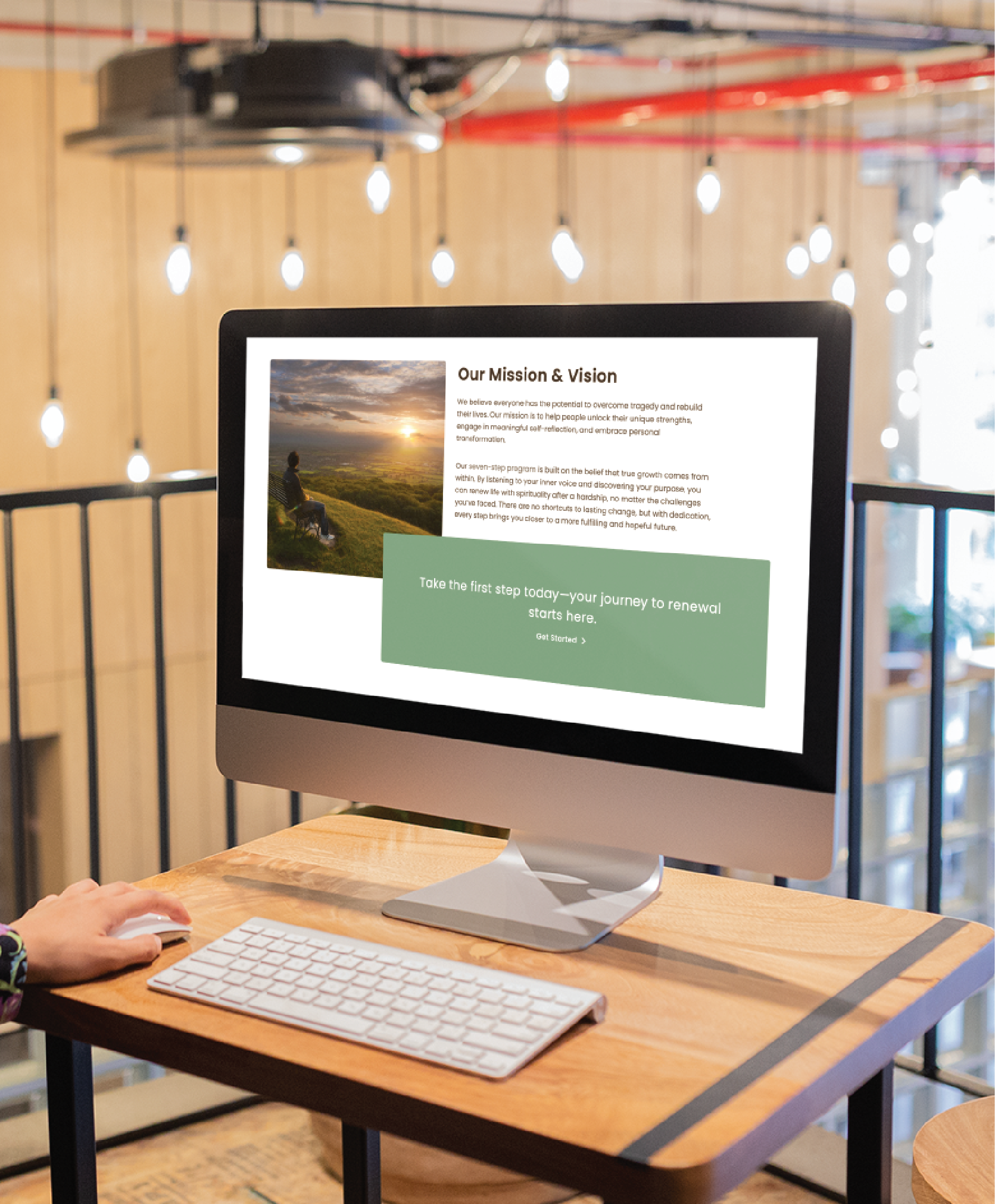A person sits at a wooden desk using a desktop computer in a modern, well-lit office. The screen showcases Professional Website Creation with mission and vision statements and a scenic photo of someone on a green hillside.