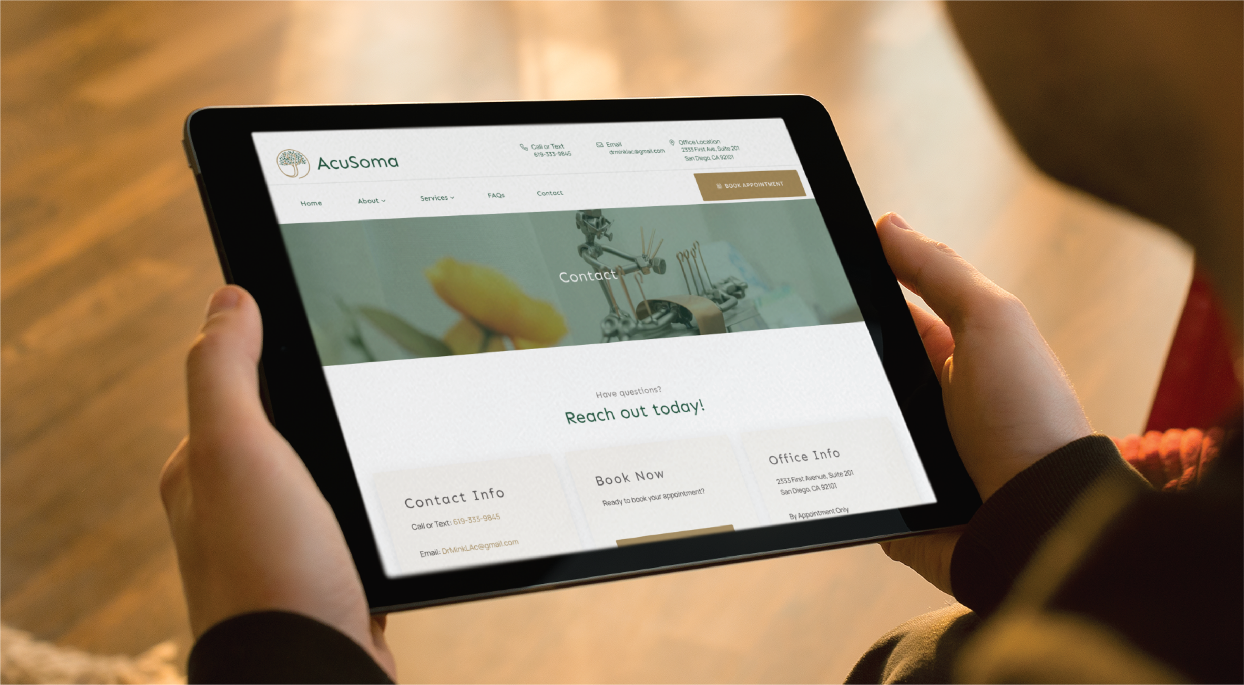 A person holds a tablet displaying the AcuSoma website’s contact page—expertly crafted through professional website creation—showing options for contact info, booking, and office info, with a blurred image of spa items in the background.
