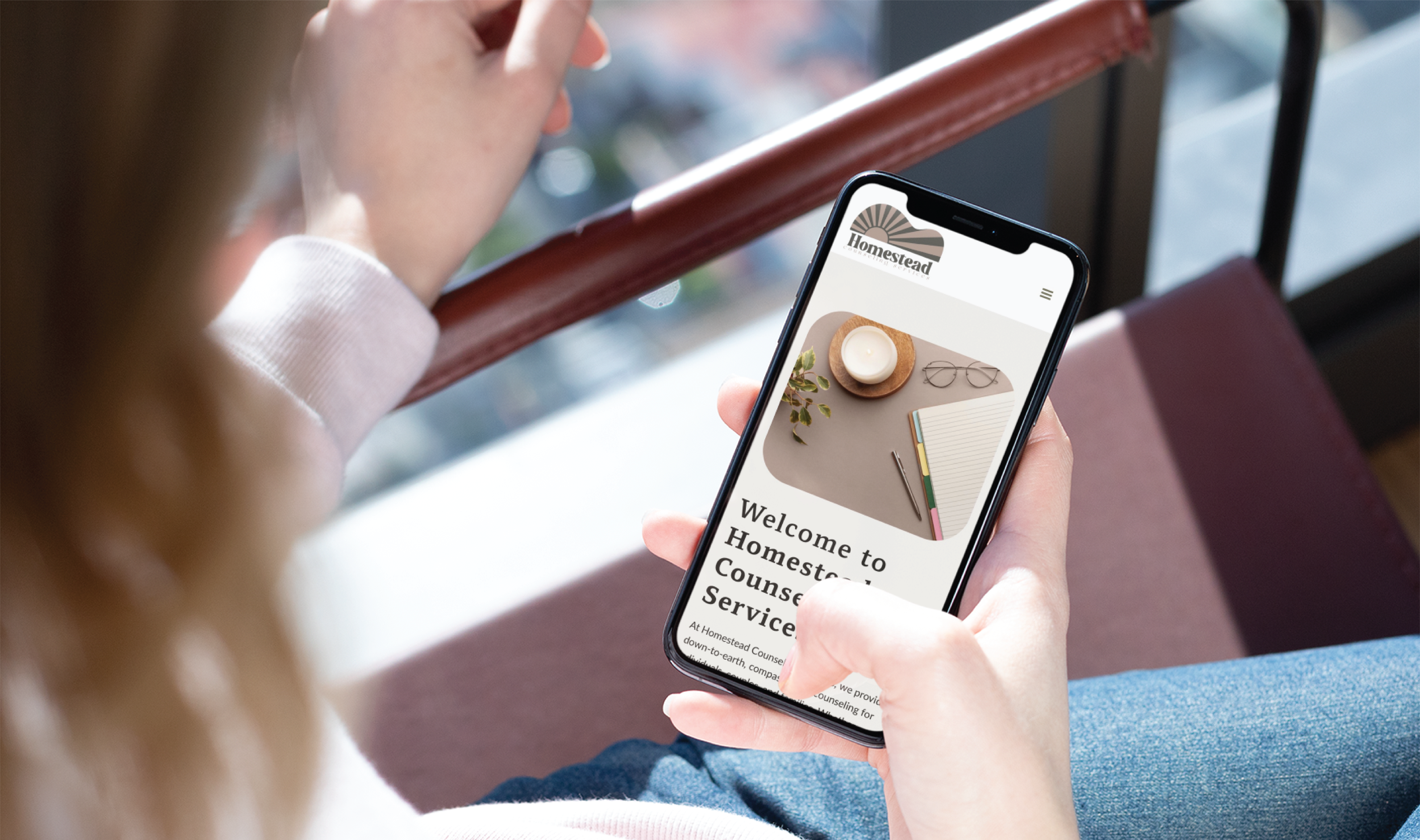 A person holding a smartphone displaying Homestead Counseling Services' website, featuring a Pennsylvania welcome message and a desk image with a candle, glasses, and stationery—showcasing thoughtful website design and organization.