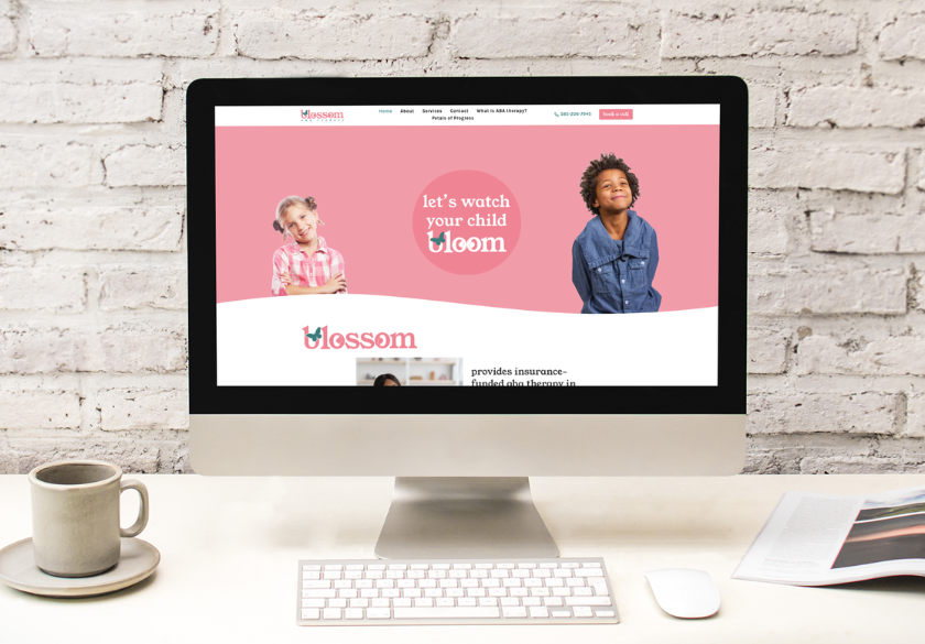 A desktop computer on a desk displays a website with photos of two smiling children and the text lets watch your child bloom, while the workspace includes a coffee cup, saucer, smartphone, papers—and no ads or website issues in sight.