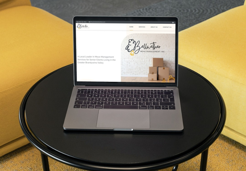 A laptop sits on a black round table, displaying a house management website that highlights how to fix website issues. Boxes and a brick wall feature in the banner, while yellow upholstered furniture is visible in the background.