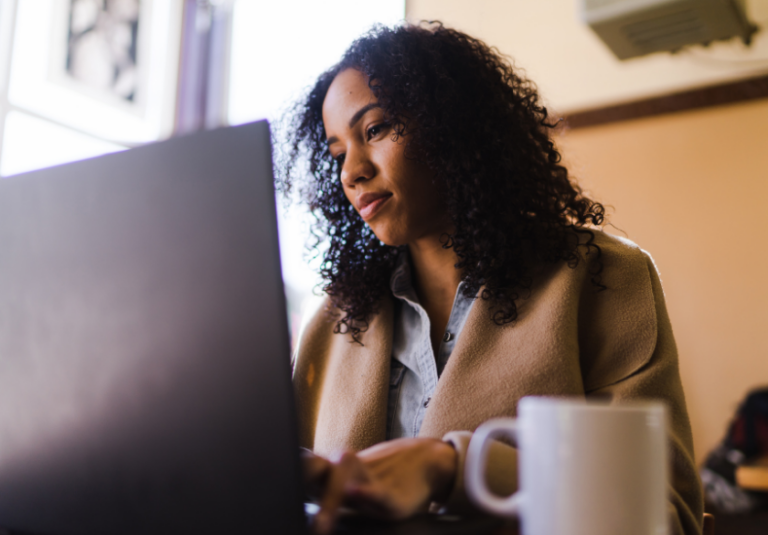 A woman with curly hair sits at a table using a laptop, wearing a light brown coat and blue shirt. A white mug is in the foreground as she appears focused on web design, likely enhancing website usability through user-centered design principles.