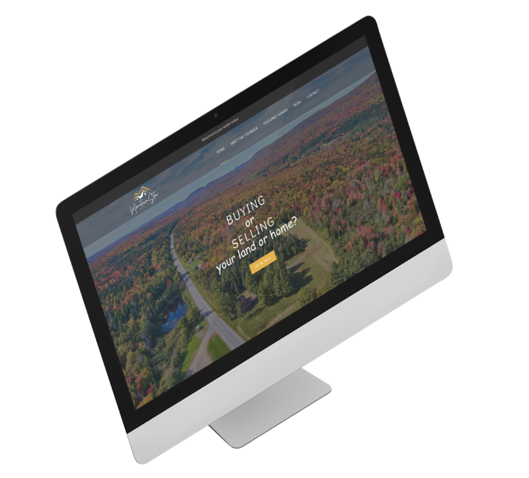 An angled desktop computer screen displays a real estate website featuring an aerial view of a forest and road. The screen shows the text Buying or selling your land or home? in white letters over the scenic background.