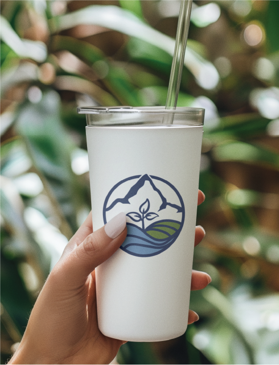 A hand holds a white reusable cup with a clear straw. The cup features a logo design of a mountain, fields, and a sprouting plant. Green foliage is blurred in the background, hinting at Kansas City's vibrant non profit community.