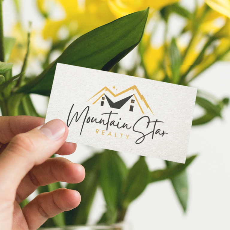 A hand holds a business card with the logo and text Mountain Star Realty, featuring a custom print design with a stylized mountain, two house icons, and yellow accents. Bright yellow flowers and green leaves are blurred in the background.