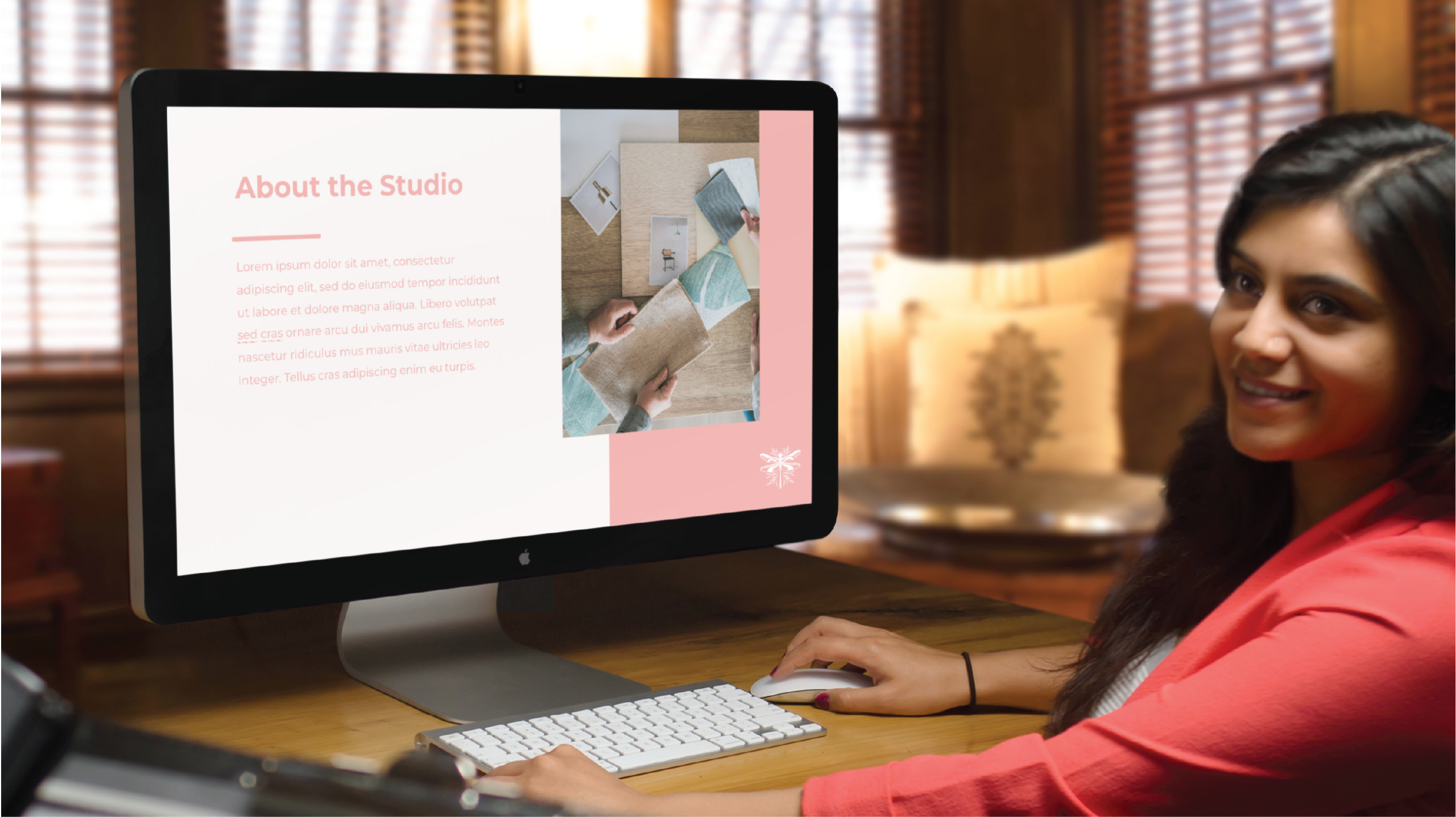 A woman in a coral blazer sits at a desk in a warmly lit Florida office with large windows and wooden blinds, working on Social Graphics as her computer displays an "About the Studio" slide with text and images.
