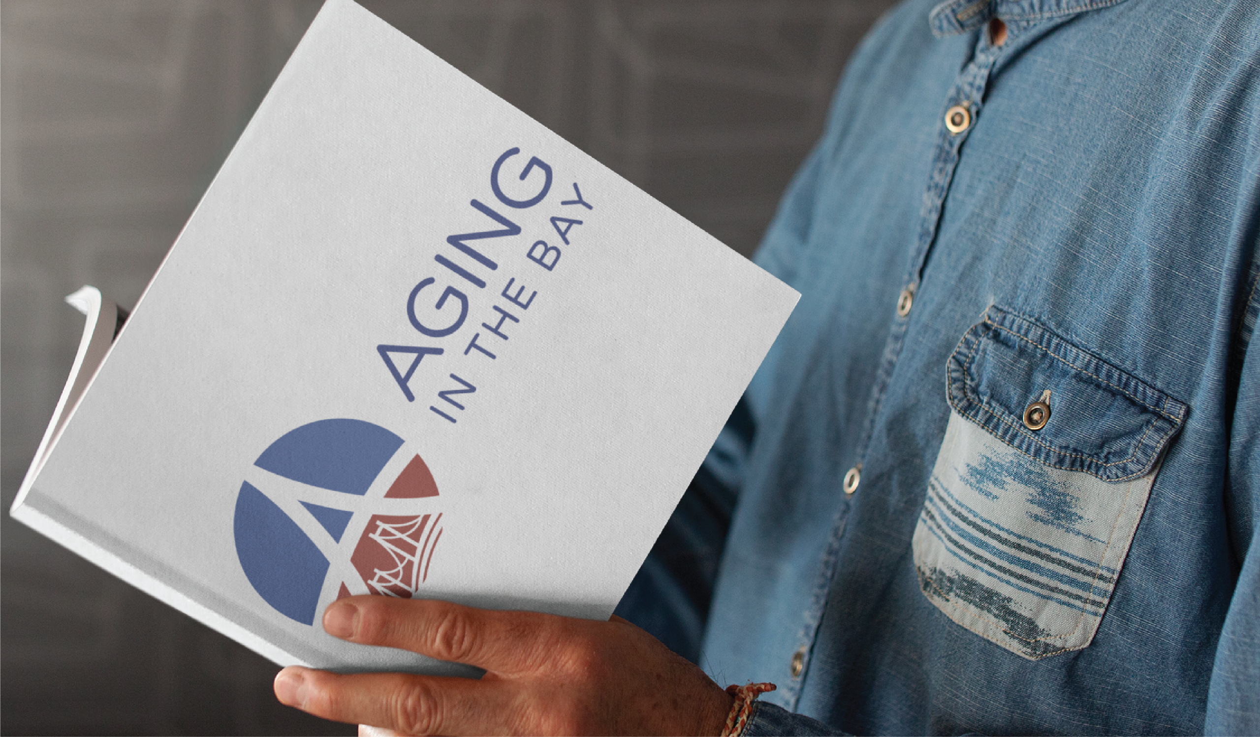 A person in a blue patterned shirt holds and reads a book titled Aging in the Bay, featuring a stylized logo design on the cover. The blurred, neutral background evokes the calm of a Montana nature preserve.