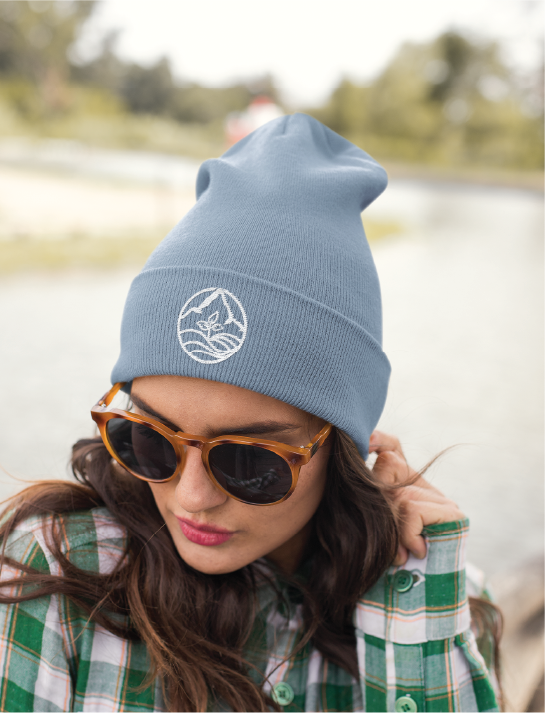 A woman wearing brown sunglasses and a blue beanie with a white logo design, dressed in a green plaid shirt, looks down while standing outdoors near water in Kansas City.