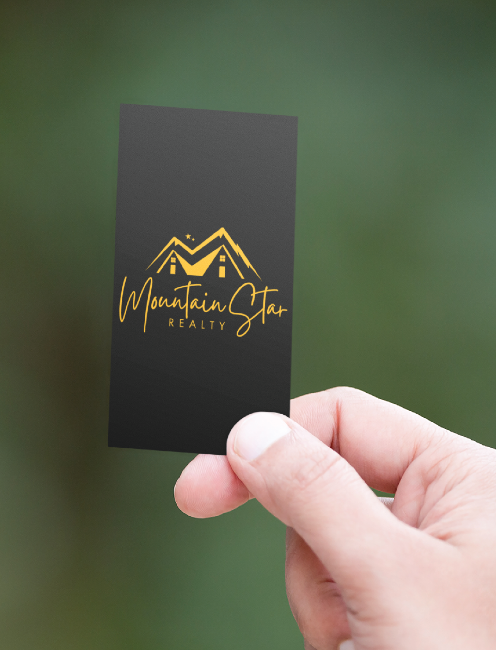 custom business card design for realty | Moonlit Media LLC A hand holds a black business card featuring a golden logo of two mountain peaks with windows, stars, and the text Mountain Star Realty in elegant handwriting and block lettering. Sleek stationery design stands out against the blurred green Michigan background.