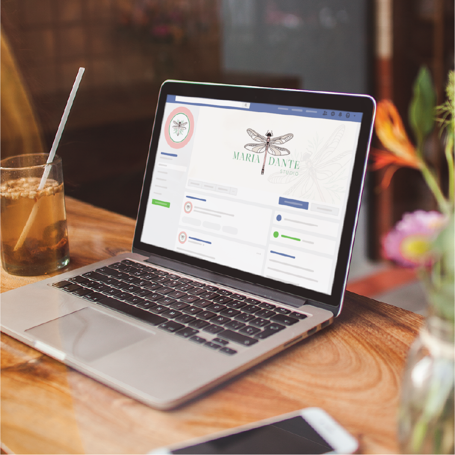A laptop on a wooden table displays a Facebook business page for Maria Dante Studio, an interior designer in Florida, with a dragonfly logo. Next to the laptop is an iced drink with a straw, vibrant flowers, and stylish social graphics on screen.