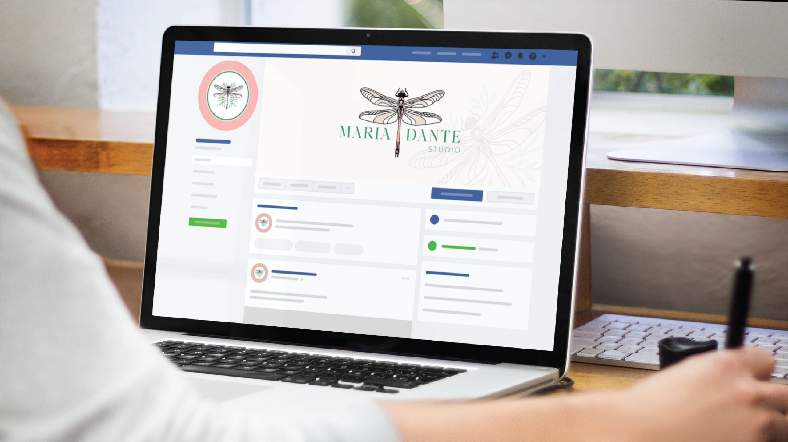 A person uses a laptop displaying a Facebook business page for Maria Dante Studio, an Interior Designer in Florida, featuring a dragonfly logo and profile image, while sitting at a desk with a notepad and pen nearby.