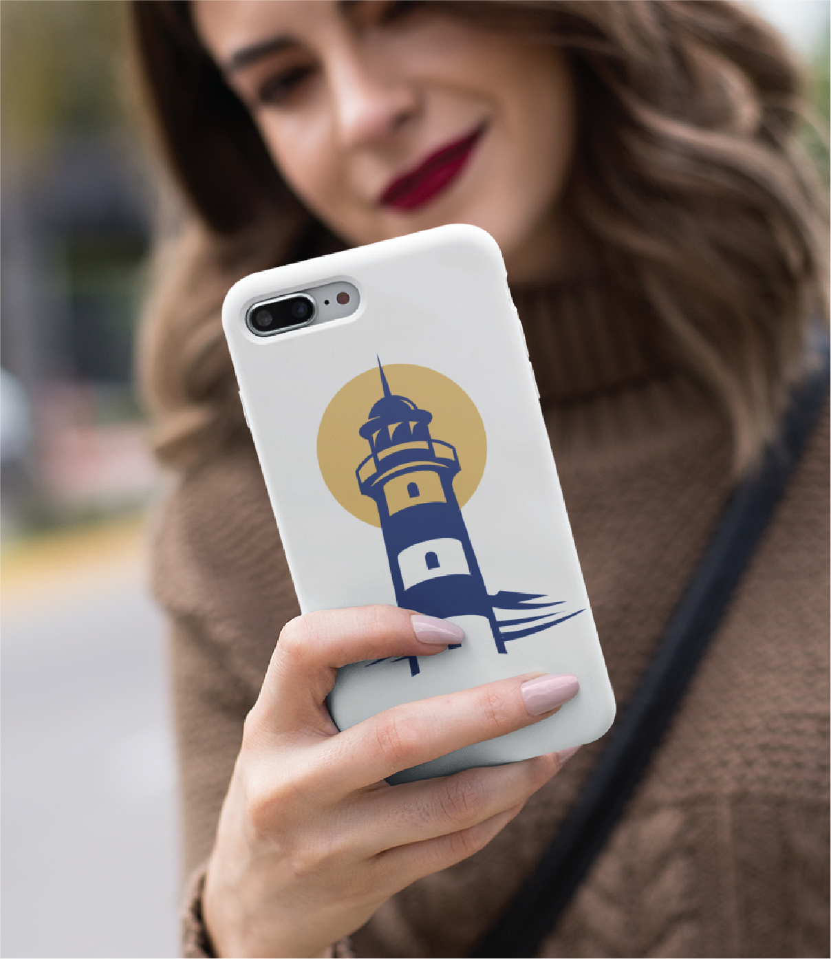 A woman in a brown sweater holds a white phone featuring a lighthouse logo design with a yellow circle, smiling as she looks at the screen outdoors in California.